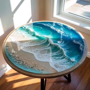 May include: Round wooden side table with a resin top depicting an ocean scene. The top features a beach with sand, pebbles, and foamy waves in shades of white, turquoise, and blue. The table has a dark wooden frame and black metal legs.