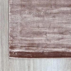 May include: A rectangular area rug in a dusty mauve or muted rose color with a subtle horizontal sheen and a soft, textured pile. The surface features a watermark text overlay that reads Rugs by Zaifa in the center of the rug's visible section.