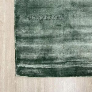 May include: A rectangular area rug in a deep forest green hue with a soft, lustrous sheen and a subtle horizontal texture. The surface features a visible watermark overlay with the text Rugs by Zaifa centered on the fabric pile.