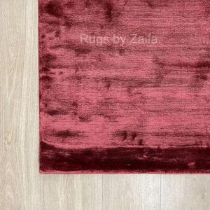 May include: A deep burgundy area rug with a soft, lustrous pile and a subtle sheen. The surface features a textured, monochromatic appearance with variations in light reflection. The text "Rugs by Zaila" is visible in the center of the rug.