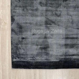 May include: A rectangular area rug featuring a dark charcoal gray and black color palette with a subtle, mottled, and horizontal striated texture. The surface displays a soft sheen and includes the white text Rugs by Zaila centered on the rug.