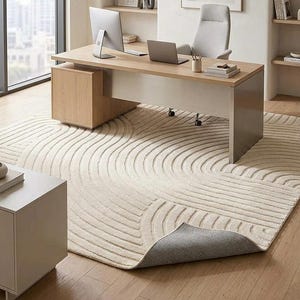 May include: A large rectangular area rug featuring a cream-colored textured surface with raised concentric semi-circular line patterns. One corner of the rug is folded over to reveal a dark gray felt backing, contrasting with the light-colored top.