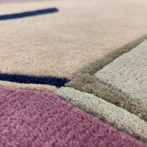 May include: A textured area rug featuring a geometric pattern with distinct color blocks in shades of dusty pink, muted beige, light gray, and deep navy blue. The surface has a dense, tufted pile construction with clearly defined, recessed borders.