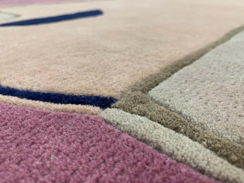 May include: A textured area rug featuring a geometric pattern with distinct color blocks in shades of dusty pink, muted beige, light gray, and deep navy blue. The surface has a dense, tufted pile construction with clearly defined, recessed borders.