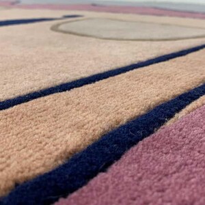 May include: A textured area rug featuring a geometric pattern with blocks of soft pink, beige, and deep navy blue. The surface has a dense, tufted pile construction with distinct color-blocked sections separated by bold, dark blue linear borders.