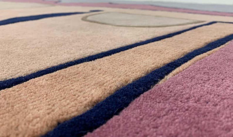 May include: A textured area rug featuring a geometric pattern with blocks of soft pink, beige, and deep navy blue. The surface has a dense, tufted pile construction with distinct color-blocked sections separated by bold, dark blue linear borders.