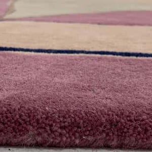 May include: A plush area rug featuring a geometric pattern with blocks of dusty rose, beige, and light gray, separated by thin, dark navy blue lines. The rug has a thick, soft pile texture and a clean, finished edge along the bottom border.