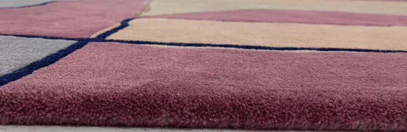 May include: A plush area rug featuring a geometric pattern with blocks of dusty rose, beige, and light gray, separated by thin, dark navy blue lines. The rug has a thick, soft pile texture and a clean, finished edge along the bottom border.