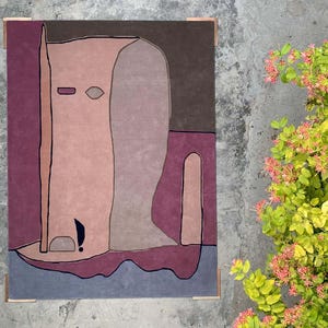 May include: A rectangular area rug featuring an abstract design with large, organic shapes in shades of dusty pink, deep plum, dark brown, and slate blue. The composition includes bold black outlines defining the various color-blocked sections.