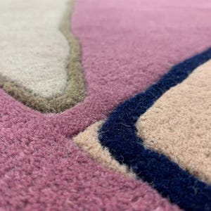 May include: A tufted rug featuring an abstract pattern of interlocking shapes in shades of mauve, beige, and cream. The sections are defined by thick, raised borders in dark navy blue and olive green, creating a textured, multi-layered surface.