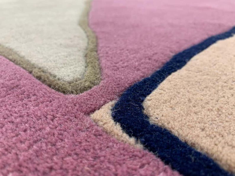 May include: A tufted rug featuring an abstract pattern of interlocking shapes in shades of mauve, beige, and cream. The sections are defined by thick, raised borders in dark navy blue and olive green, creating a textured, multi-layered surface.