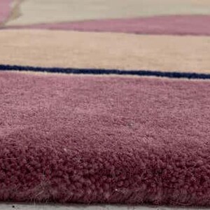 May include: A plush area rug featuring a geometric pattern with blocks of dusty rose, beige, and light gray, separated by thin, dark navy blue lines. The rug has a thick, soft pile texture and a clean, finished edge along the bottom border.