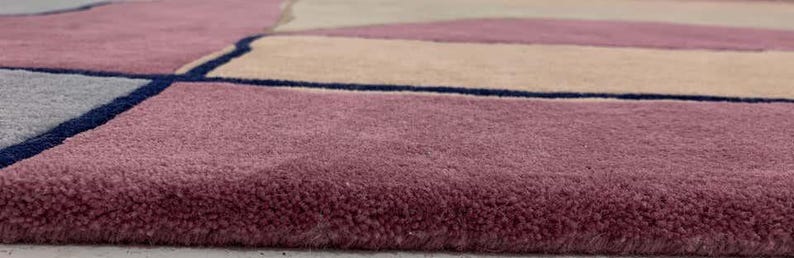 May include: A plush area rug featuring a geometric pattern with blocks of dusty rose, beige, and light gray, separated by thin, dark navy blue lines. The rug has a thick, soft pile texture and a clean, finished edge along the bottom border.