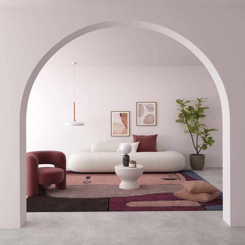 May include: A living room interior featuring a curved white sofa, a dark red armchair, and a large area rug with abstract shapes in shades of pink, purple, and blue. A white pedestal table, a potted plant, and two framed wall art pieces are included.