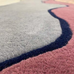 May include: A plush area rug featuring a soft gray section and a dusty rose section, separated by a distinct, dark navy blue wavy border. The rug has a thick, tufted texture with clearly defined color blocks and a smooth, consistent pile height.