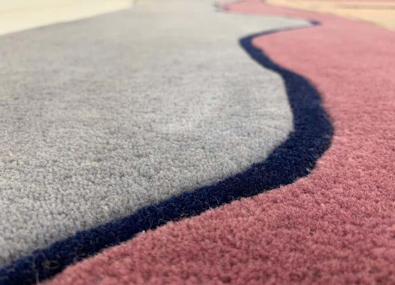 May include: A plush area rug featuring a soft gray section and a dusty rose section, separated by a distinct, dark navy blue wavy border. The rug has a thick, tufted texture with clearly defined color blocks and a smooth, consistent pile height.