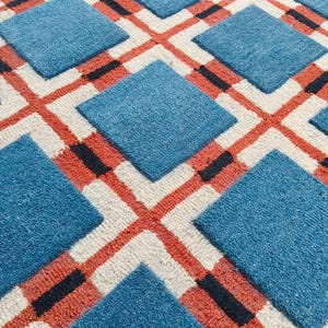 May include: A tufted rug featuring a repeating geometric pattern of solid blue squares arranged in a grid. The squares are separated by intersecting lines of cream-colored yarn, accented with thin, horizontal stripes of orange and black.