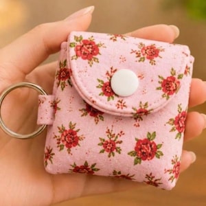 Può includere: Una piccola custodia quadrata in tessuto rosa chiaro con un motivo ripetuto di rose rosse e foglie verdi. La custodia presenta una patta arrotondata chiusa da un bottone a pressione in plastica bianca e un anello portachiavi in metallo argentato.