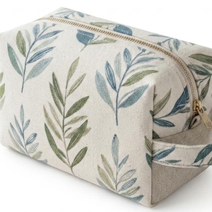 May include: A rectangular toiletry bag made of off-white fabric featuring a repeating pattern of green and blue leaves. The bag includes a gold-toned metal zipper closure along the top and a matching fabric handle attached to the side panel.