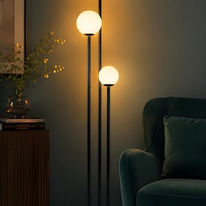May include: A floor lamp featuring a slender black metal frame with three vertical poles of varying heights. Each pole is topped with a spherical white glass globe light, emitting a warm glow. The lamp stands on a circular black metal base.