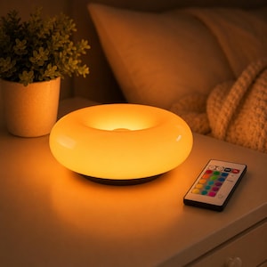 Peut inclure: Une lampe de table en forme de beignet diffusant une lueur orange chaude, dotée d'une surface lisse et arrondie. À côté de la lampe se trouve une petite télécommande rectangulaire avec plusieurs boutons colorés pour régler la lumière.