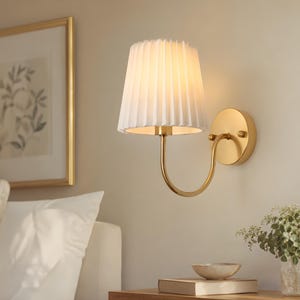 Peut inclure: Une applique murale composée d'un bras incurvé en laiton et d'une plaque de base circulaire. La lampe est surmontée d'un abat-jour en tissu blanc plissé de forme conique qui diffuse une lumière chaude depuis la structure du luminaire.