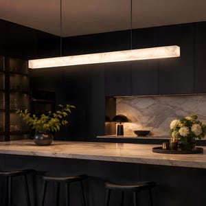 May include: A long, rectangular linear pendant light fixture with a translucent, marbled white surface that emits a warm, soft glow. The fixture is suspended by two thin, dark wires from the ceiling, positioned horizontally above a kitchen island.