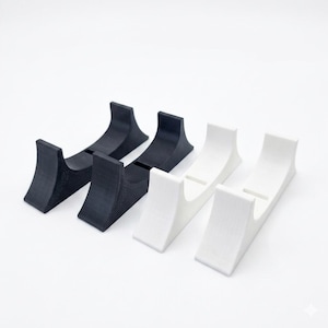 Puede incluir: Dos soportes negros y dos blancos curvados diseñados para sostener objetos alargados. Cada soporte presenta una superficie superior cóncava con un recorte rectangular central, proporcionando una base estable para apoyar objetos horizontalmente.