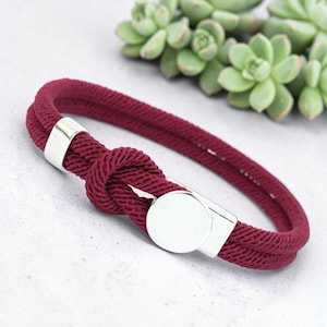 May include: A burgundy rope bracelet with a knot design and silver-colored metal clasp. The bracelet is made of a thick, woven material. The clasp is a circular shape. The background includes green succulent plants.
