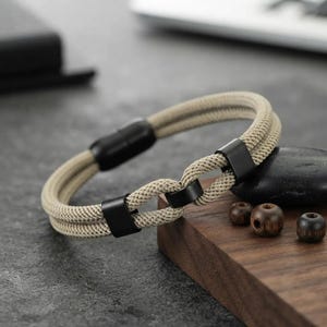 Puede incluir: Una pulsera de cuerda beige de doble hebra con tres conectores rectangulares de metal negro mate y un cierre magnético cilíndrico negro a juego. La pulsera está fabricada con cordón trenzado e incluye un mecanismo de cierre elegante.
