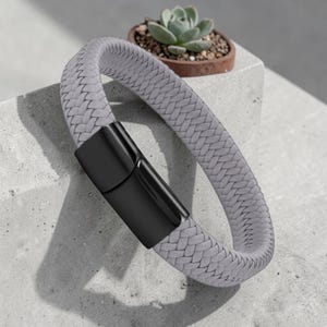 May include: A gray braided bracelet with a black magnetic clasp. The bracelet is made of a woven material and is displayed on a concrete surface. A small potted succulent is in the background.