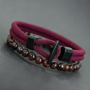 Könnte beinhalten: Zwei Armbänder. Das obere Armband ist ein tiefrotes Seildesign mit schwarzen Metallakzenten. Das untere Armband hat dunkelbraune, runde Perlen. Die Armbänder werden auf einer dunkelgrauen Oberfläche präsentiert.