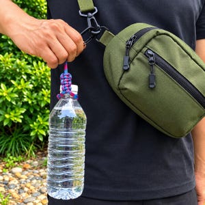 Puede incluir: Un bolso bandolera de color verde oliva con cremallera negra y correa a juego. Una botella de agua de plástico transparente está sujeta a la hebilla del bolso mediante un soporte de paracord trenzado con patrones en azul, rosa y morado.