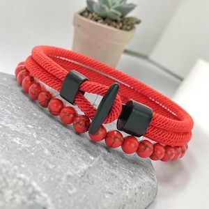 Op de afbeelding: Een rode armband met een geweven touwontwerp en een rij rode kralen. De armband heeft zwarte sluitingen en een tuimelsluiting. De kralen hebben een gemarmerd patroon. De armband ligt op een grijs stenen oppervlak.