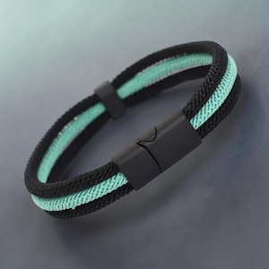 Könnte beinhalten: Ein schwarzes und mintgrünes Armband mit Magnetverschluss. Das Armband hat ein gewebtes Design mit einer schwarzen Außenschicht und einer mintgrünen Innenschicht. Der Verschluss ist schwarz und rechteckig. Ein stilvolles Accessoire.