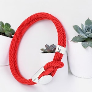 Op de afbeelding: Een felrode touw armband met een zilveren sluiting. De armband is gelust en vastgemaakt met een rechthoekige en ovale zilveren sluiting. De achtergrond toont witte potten met groene vetplanten.