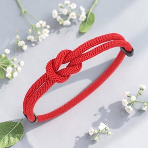 May include: A red rope bracelet with a knot design. The bracelet is made of a thick, woven material and features a knot in the center. The bracelet is a bright red color and has black end pieces.