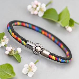 May include: A braided bracelet with a black band and a rainbow-colored inner design. The bracelet features a silver-colored clasp with a decorative pattern. The jewelry is displayed on a light gray surface with white flowers and green leaves.