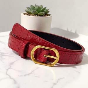 Op de afbeelding: Een rode riem met een glanzende krokodil-reliëftextuur en een goudkleurige ovale gesp. De riem is voorzien van een bijpassende rode lus en een zwarte binnenvoering, met zichtbare stiksels langs de randen van de riem.