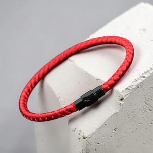 Op de afbeelding: Een rode gevlochten armband met een zwarte sluiting. De armband is cirkelvormig en lijkt gemaakt van een geweven materiaal. De sluiting is rechthoekig en verbindt de twee uiteinden van de armband.