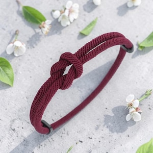 Könnte beinhalten: Ein bordeauxfarbenes Seilarmband mit einem geknoteten Design. Das Armband besteht aus einem dicken, strukturierten Seil. Der Hintergrund umfasst weiße Blumen und grüne Blätter. Der Stil des Armbands ist schlicht und elegant.