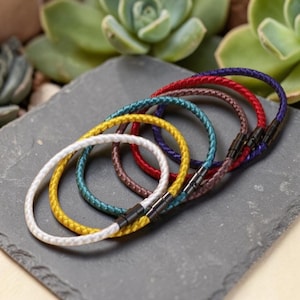 Op de afbeelding: Een verzameling gevlochten armbanden in verschillende kleuren: wit, geel, turquoise, bruin, rood en paars. Elke armband heeft een magnetische sluiting. De armbanden liggen op een grijze stenen ondergrond, met groene planten op de achtergrond.