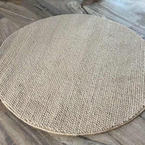 May include: A round, beige rug with a textured surface. The rug is made of small, tightly packed, raised loops, creating a nubby appearance. The rug is placed on a light-colored, patterned floor.