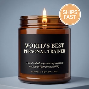 May include: A lit candle in an amber glass jar with a black label. The label reads: WORLD'S BEST PERSONAL TRAINER, A sweat-salted, rep-counting aroma of 100% gym-floor accountability, NOVARI | SOY WAX 9OZ. The candle has a visible flame at the top.
