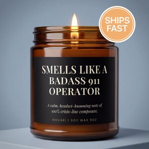 May include: A lit brown glass candle jar with a black label that reads "SMELLS LIKE A BADASS 911 OPERATOR." The candle has a "SHIPS FAST" sticker. The text below the title reads "A calm, headset-humming note of 100% crisis-line composure."