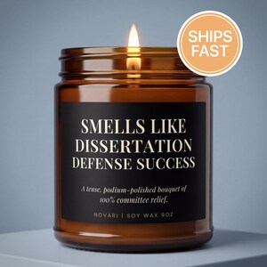 May include: A lit brown glass jar candle with a black label that reads "SMELLS LIKE DISSERTATION DEFENSE SUCCESS." The candle also says "A tense, podium-polished bouquet of 100% committee relief." and "NOVARI | SOY WAX 9OZ." A "SHIPS FAST" sticker is on the top.