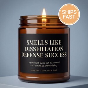 May include: A lit soy wax candle in an amber glass jar with a black label. The label reads: SMELLS LIKE DISSERTATION DEFENSE SUCCESS. A parchment-warm, oak-lit aroma of 100% committee approved glory. NOVARI SOY WAX 9OZ. A flame burns at the top.