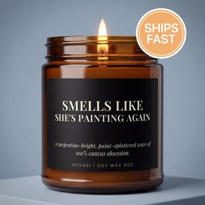 May include: A lit soy wax candle in an amber glass jar with a black label. The text reads: SMELLS LIKE SHE'S PAINTING AGAIN, A turpentine-bright, paint-splattered wave of 100% canvas obsession. NOVARI | SOY WAX 9OZ. The candle has a single burning wick.