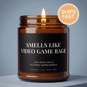 May include: A lit candle in an amber glass jar with a black label that reads: SMELLS LIKE VIDEO GAME RAGE, A hot, chaotic aroma of 100% button-mashing meltdown. NOVARI SOY WAX 9OZ. The candle features a visible flame and a warm, glowing wax surface.