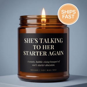 May include: A lit brown glass jar candle with the text "SHE'S TALKING TO HER STARTER AGAIN" on a black label. The candle has a "SHIPS FAST" sticker. The candle is 9 oz and made of soy wax.